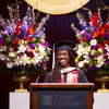 A graduation speaker in a cap and gown smiling at a podium with flowers in the background